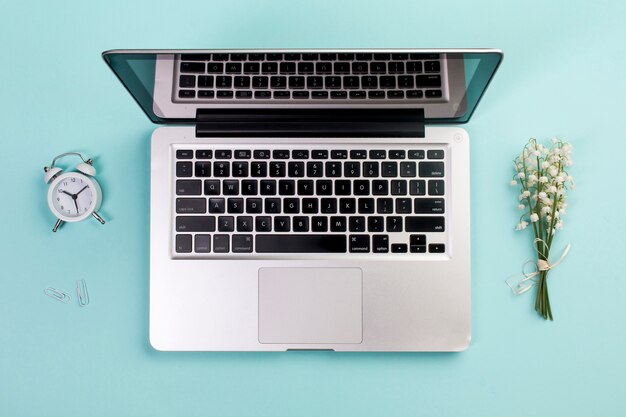 Réveil, trombone, bouquet de muguet avec un ordinateur portable ouvert sur le bureau bleu