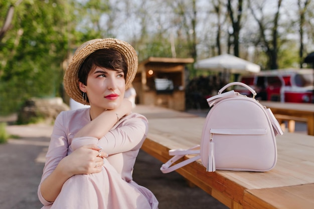 Rêver de jeune femme aux cheveux bruns brillants en détournant les yeux, en attendant la commande dans un restaurant en plein air. Portrait de jolie fille brune en tenue tendance avec sac à dos en cuir allongé sur la table.