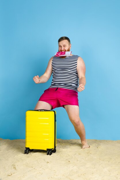 Les rêves deviennent réalité. Heureux jeune homme avec sac préparé pour voyager sur fond bleu studio. Concept d'émotions humaines, expression faciale, vacances d'été, week-end. L'été, la mer, l'océan, l'alcool.