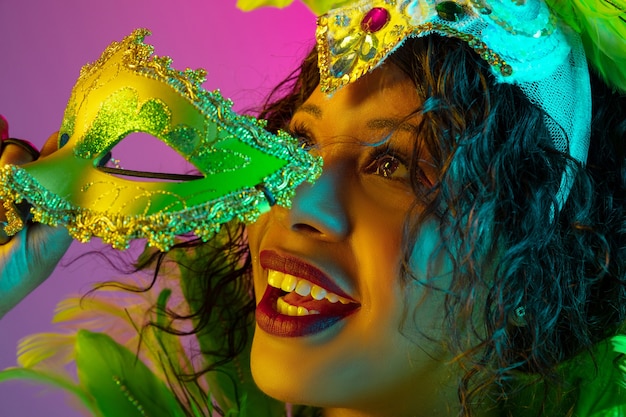 Rêveur. Belle jeune femme en carnaval, costume de mascarade élégant avec des plumes dansant sur un mur dégradé en néon. Concept de célébration de vacances, temps festif, danse, fête, s'amuser.