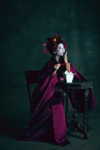 Rêveur. Jeune femme japonaise comme geisha isolée sur un mur vert foncé. Style rétro, comparaison du concept d'époques. Beau modèle féminin comme personnage historique brillant, démodé.