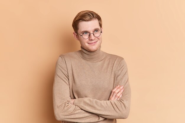 Rêveur satisfait aux yeux bleus beau jeune homme se tient les mains croisées à l'intérieur regarde à distance pense à quelque chose d'agréable porte un col roulé beige décontracté