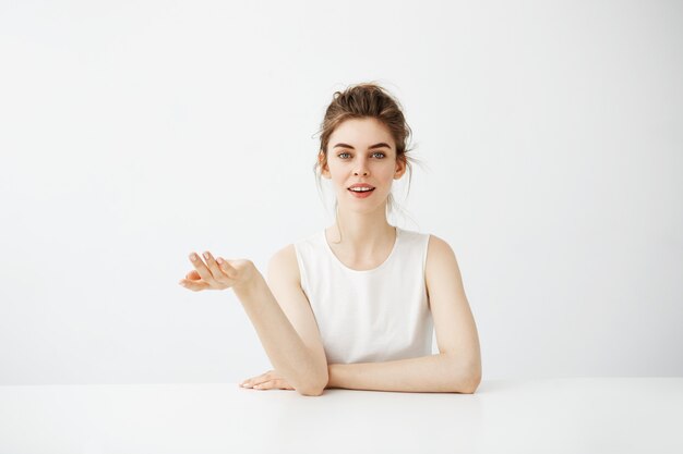 Rêveuse belle jeune femme avec chignon assis à table sur fond blanc pensant rêver.