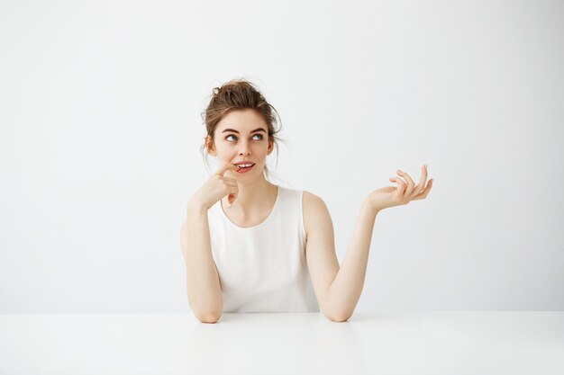 Rêveuse belle jeune femme avec chignon assis à table sur fond blanc pensant rêver.