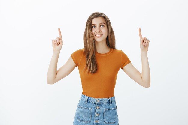 Rêveuse belle jeune femme pointant les doigts vers le haut et à la réflexion, faire un choix, souriant tout en décidant