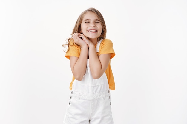 Photo gratuite rêveuse joyeuse petite fille aux cheveux blonds, serrer les mains ensemble avec joie ne peut pas attendre un événement exceptionnel, fermer les yeux souriant rêveur, se sentir ravie et heureuse, tenir un mur blanc