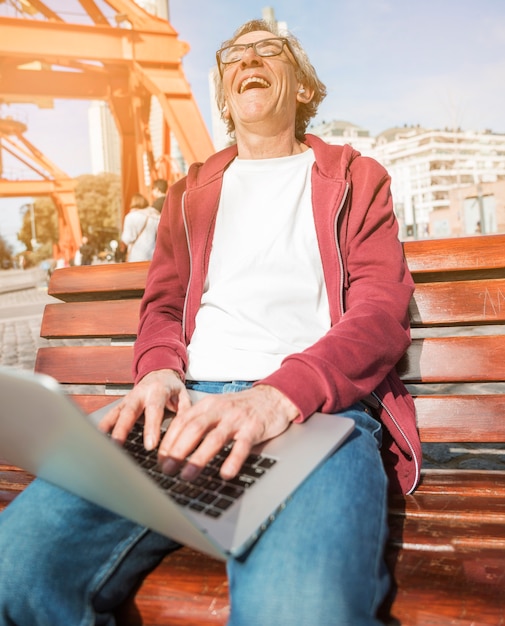 Riant senior homme assis sur un banc avec un ordinateur portable ouvert sur ses genoux