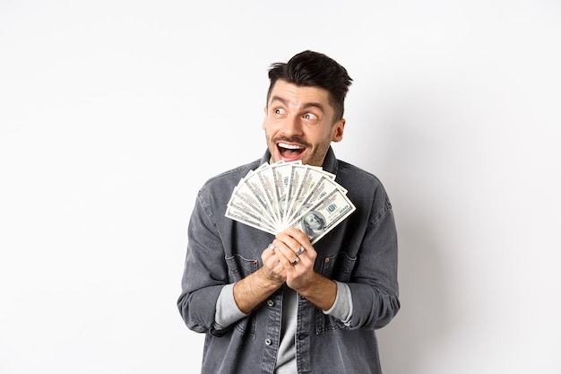 Riche homme heureux rêvant de faire du shopping souhaitant des billets d'un dollar dans les mains regardant de côté pensif et tenant de l'argent debout sur fond blanc