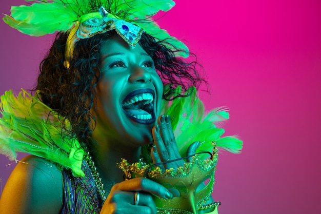 Rire. Belle jeune femme en carnaval, costume de mascarade élégant avec des plumes dansant sur un mur dégradé en néon. Concept de célébration de vacances, temps festif, danse, fête, s'amuser.