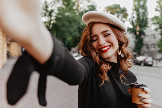 Rire charmante femme avec un maquillage lumineux faisant selfie en automne. Heureuse fille française bouclée buvant du café dans la rue.