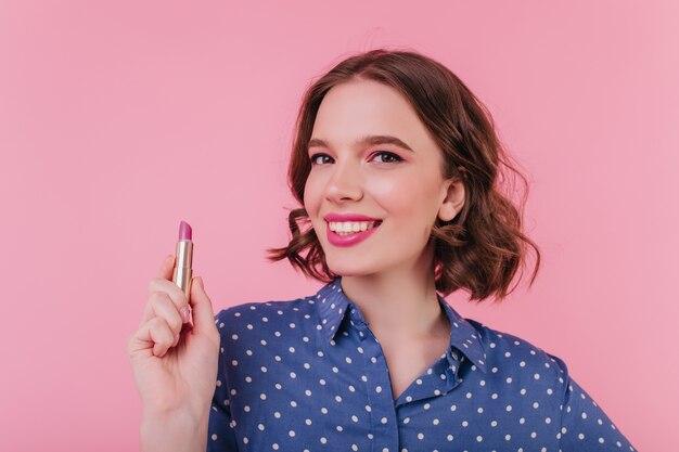 Rire de femme caucasienne aux cheveux noirs tenant le rouge à lèvres rose. Plan intérieur d'une fille extatique de bonne humeur souriant tout en faisant du maquillage