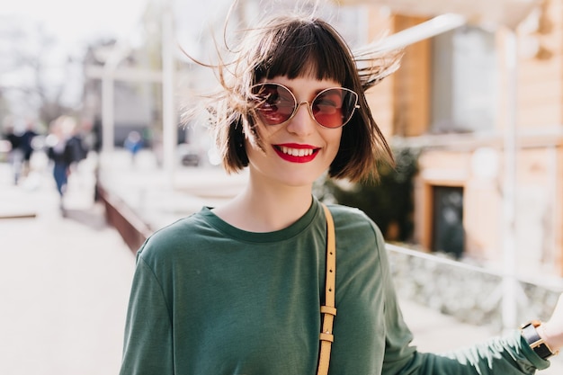 Rire femme joyeuse en lunettes de soleil marron posant sur fond de rue Photo en plein air d'une dame européenne heureuse en pull vert