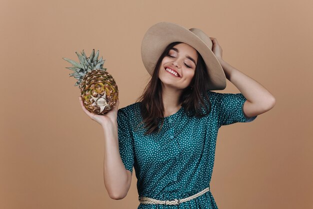 Rire fille au chapeau et robe verte pose avec un ananas dans le studio