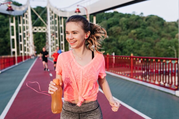 Rire fille blonde tenant une bouteille de jus et courir sur la piste de cendre. Incroyable modèle féminin s'amusant au stade.