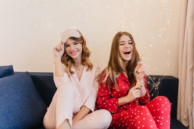 Photo gratuite rire fille brune jouant avec des ampoules. photo intérieure de deux dames en pyjama élégant assis sur un canapé bleu.