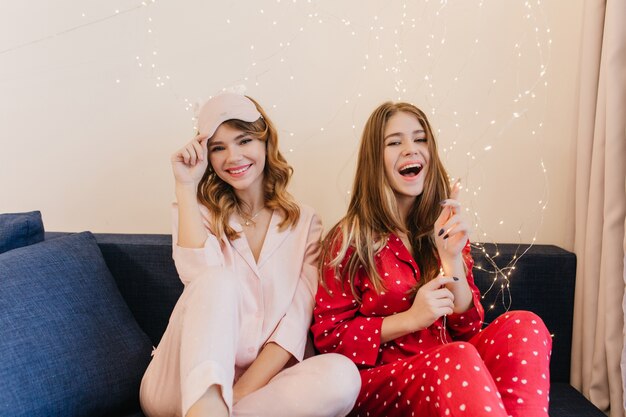 Rire fille brune jouant avec des ampoules. Photo intérieure de deux dames en pyjama élégant assis sur un canapé bleu.