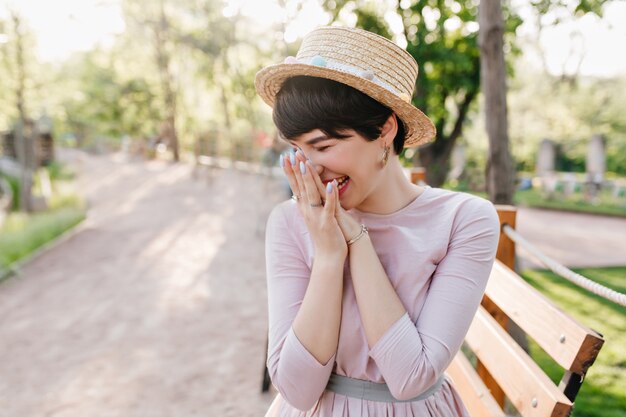 Rire fille brune à la peau pâle portant des bijoux à la mode assis sur un banc en bois dans le parc, profitant du matin ensoleillé