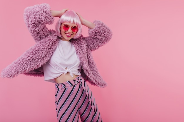 Rire fille rêveuse à lunettes de soleil touchant sa perruque brillante. Sourire incroyable jeune femme en veste de fourrure dansant sur un mur rose