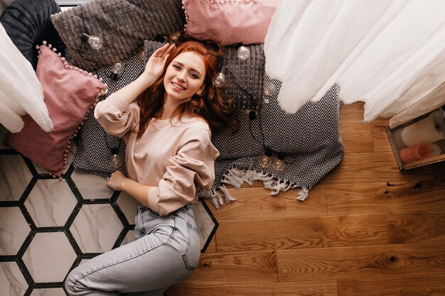 Rire fille rousse en jeans assis à côté d'oreillers. Photo intérieure d'une dame souriante au gingembre allongée sur le sol.