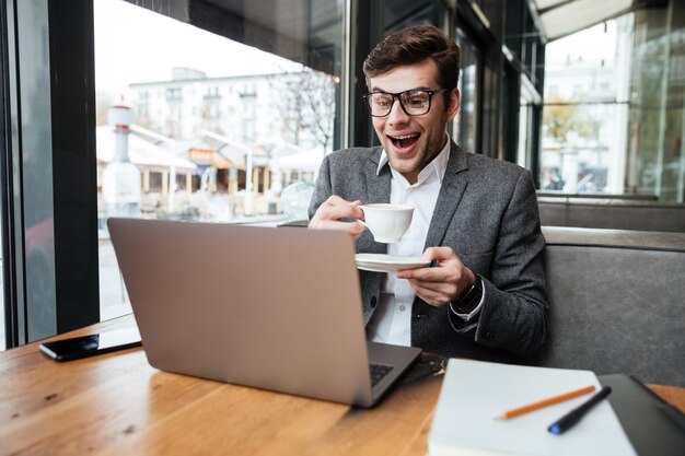 Rire homme d'affaires à lunettes assis près de la table au café avec une tasse de café tout en regardant un ordinateur portable