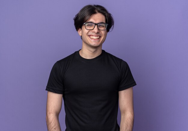 Rire jeune beau mec portant un t-shirt noir et des lunettes isolés sur un mur violet