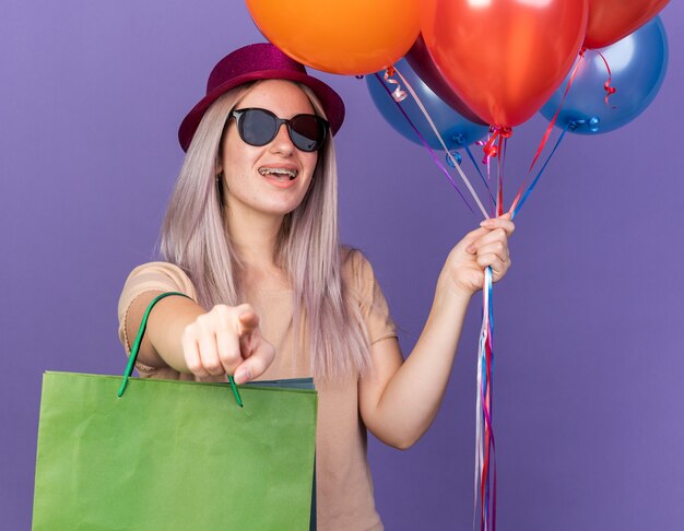 Rire jeune belle fille portant un appareil dentaire et un chapeau de fête avec des lunettes tenant des ballons avec un sac-cadeau vous montrant un geste isolé sur un mur bleu