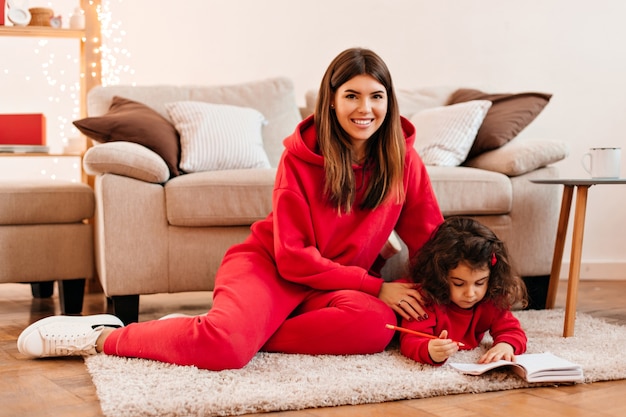 Rire jeune femme enseignant à sa fille à écrire. Plan intérieur d'une mère heureuse et d'un enfant assis sur un tapis.