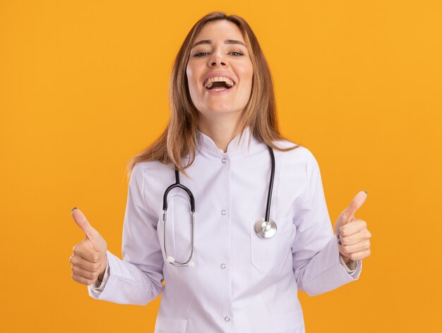 Rire jeune femme médecin portant une robe médicale avec stéthoscope montrant les pouces vers le haut isolé sur mur jaune