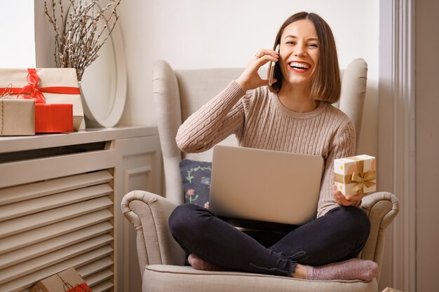 Rire jeune femme avec ordinateur portable, parler au téléphone mobile, assis dans un fauteuil confortable