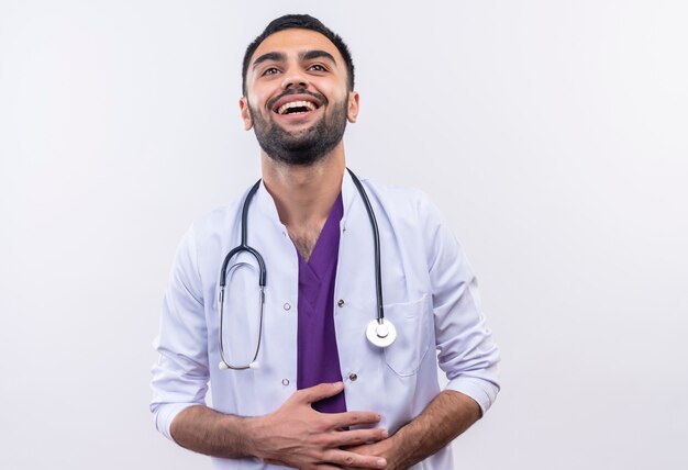 Rire jeune médecin de sexe masculin portant une robe médicale stéthoscope saisi l'estomac sur un mur blanc isolé