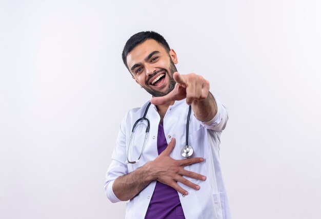 Rire jeune médecin de sexe masculin portant une robe médicale stéthoscope vous montrant le geste sur un mur blanc isolé