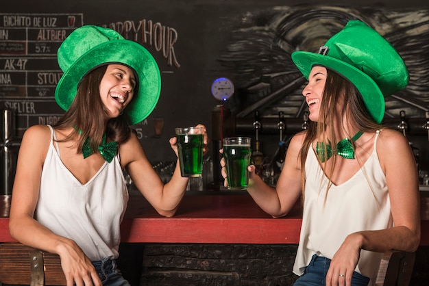 Rire de jeunes femmes en chapeaux Saint Patricks tenant des verres de boisson au comptoir