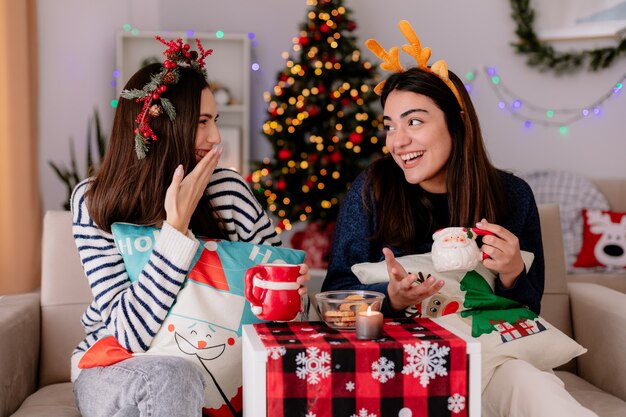 Rire de jolies jeunes filles tenir des tasses et se regarder assis sur des fauteuils et profiter du temps de Noël à la maison