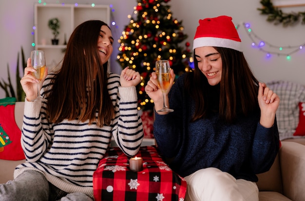 Rire de jolies jeunes filles tiennent des verres de champagne assis sur des fauteuils et profitant du temps de Noël à la maison