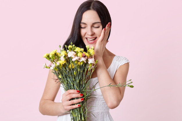 Rire séduisante jeune mannequin brune aux yeux fermés tenant de belles fleurs dans une main, touchant son visage avec l'autre