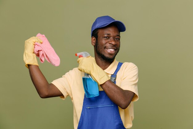 Rire tenant un agent de nettoyage avec un chiffon jeune homme nettoyant afro-américain en uniforme avec des gants isolés sur fond vert