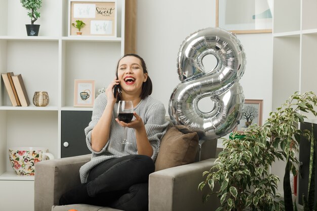 Rire les yeux fermés belle femme le jour de la femme heureuse tenant un verre de vin parle de vin assis sur un fauteuil dans le salon