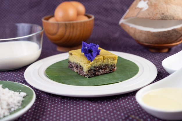 Riz gluant noir et crème anglaise sur une feuille de bananier dans une assiette blanche avec des fleurs de pois papillon.