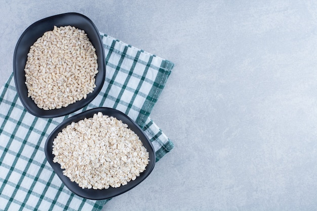 Riz à grain court et avoine empilés dans deux bols noirs sur une nappe pliée sur fond de marbre.