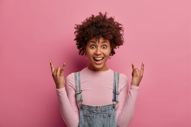 Le rock n roll est toujours vivant. Une fille à la peau sombre positive lève les mains avec un geste cool, écoute de la musique au concert, porte un col roulé et un sarafan en denim, se sent heureuse et excitée, isolée sur un mur rose