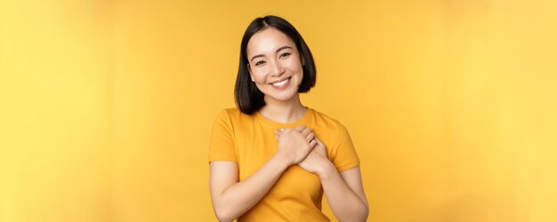 Romantique fille coréenne femme asiatique tenant les mains sur le cœur souriant avec soin et tendresse debout ov