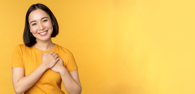 Romantique fille coréenne femme asiatique tenant les mains sur le cœur souriant avec soin et tendresse debout vo