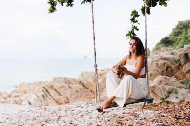 Romantique jeune femme caucasienne calme heureux avec ukulélé sur la plage rocheuse tropicale au coucher du soleil