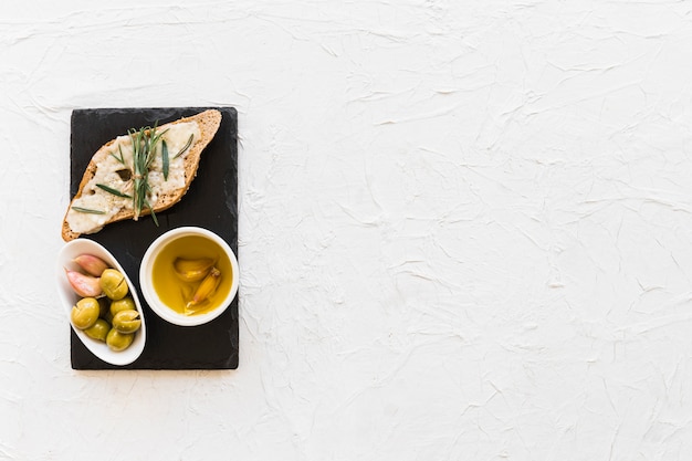Photo gratuite romarin et fromage sur le pain avec de l'huile et des olives dans le bol sur la plaque d'ardoise