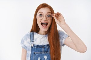 Une rouquine animée impressionnée une fille heureuse porte une salopette en jean souriant étonnée des lunettes tactiles joyeuses regarde la caméra étonnée et ravie expriment l'étonnement et la joie du kiosque à journaux cool fond blanc