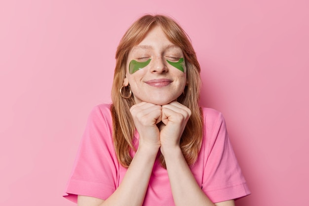 Une rouquine heureuse garde les mains sous le menton avec les yeux fermés applique des patchs d'hydrogel pour les soins de la peau porte un t-shirt décontracté isolé sur fond rose Les jeunes et les procédures de beauté