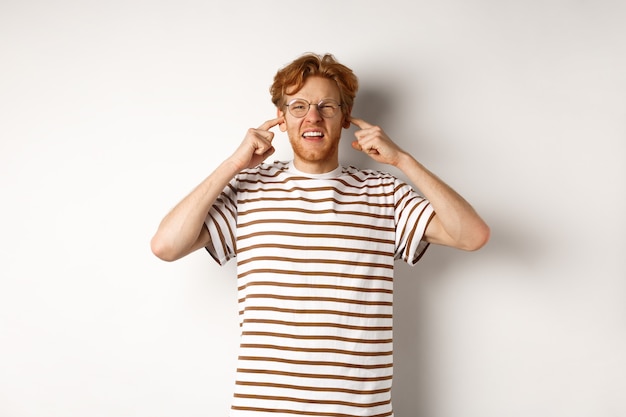 Rousse agacé à lunettes fermant les oreilles et se plaignant d'un bruit fort, en colère contre les voisins bruyants, debout sur fond blanc.