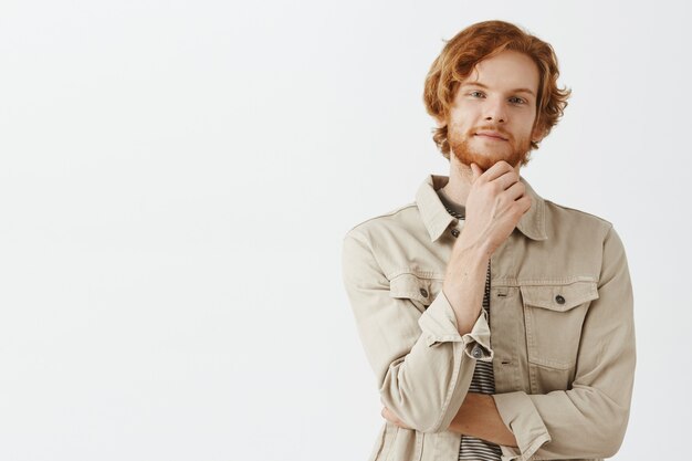 Rousse barbu réfléchie et intriguée posant contre le mur blanc