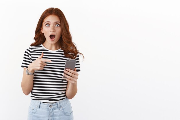 Rousse choquée et étonnée en t-shirt rayé, haletant de crainte et d'étonnement, pointant l'écran du smartphone, la caméra à la mâchoire tombante étonnée, regarde avec crainte et choc