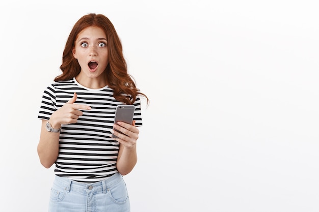 Rousse choquée et étonnée en t-shirt rayé, haletant de crainte et d'étonnement, pointant l'écran du smartphone, la caméra à la mâchoire tombante étonnée, regarde avec crainte et choc